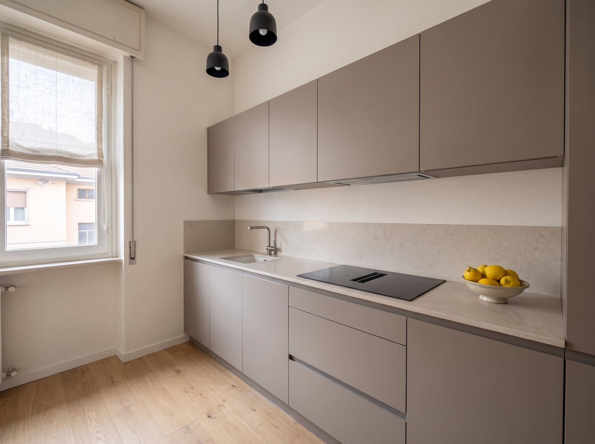 La stessa cucina dopo i lavori: mobili grigio caldo senza maniglie, piano in pietra chiara, piano a induzione, parquet chiaro.