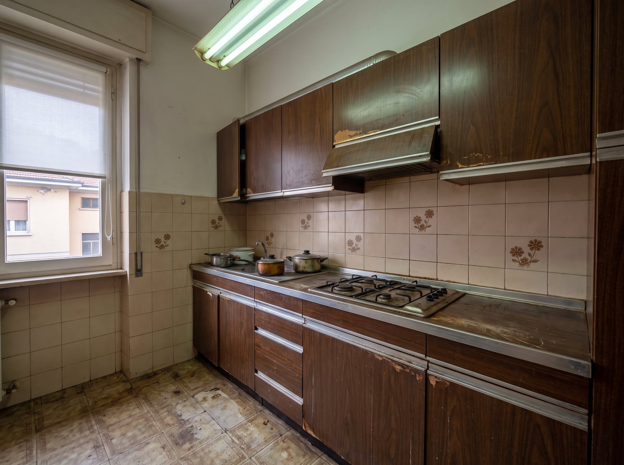 Cucina prima dei lavori: mobili in legno scuro rovinati, piastrelle beige con fiori, linoleum consumato, tubo al neon.
