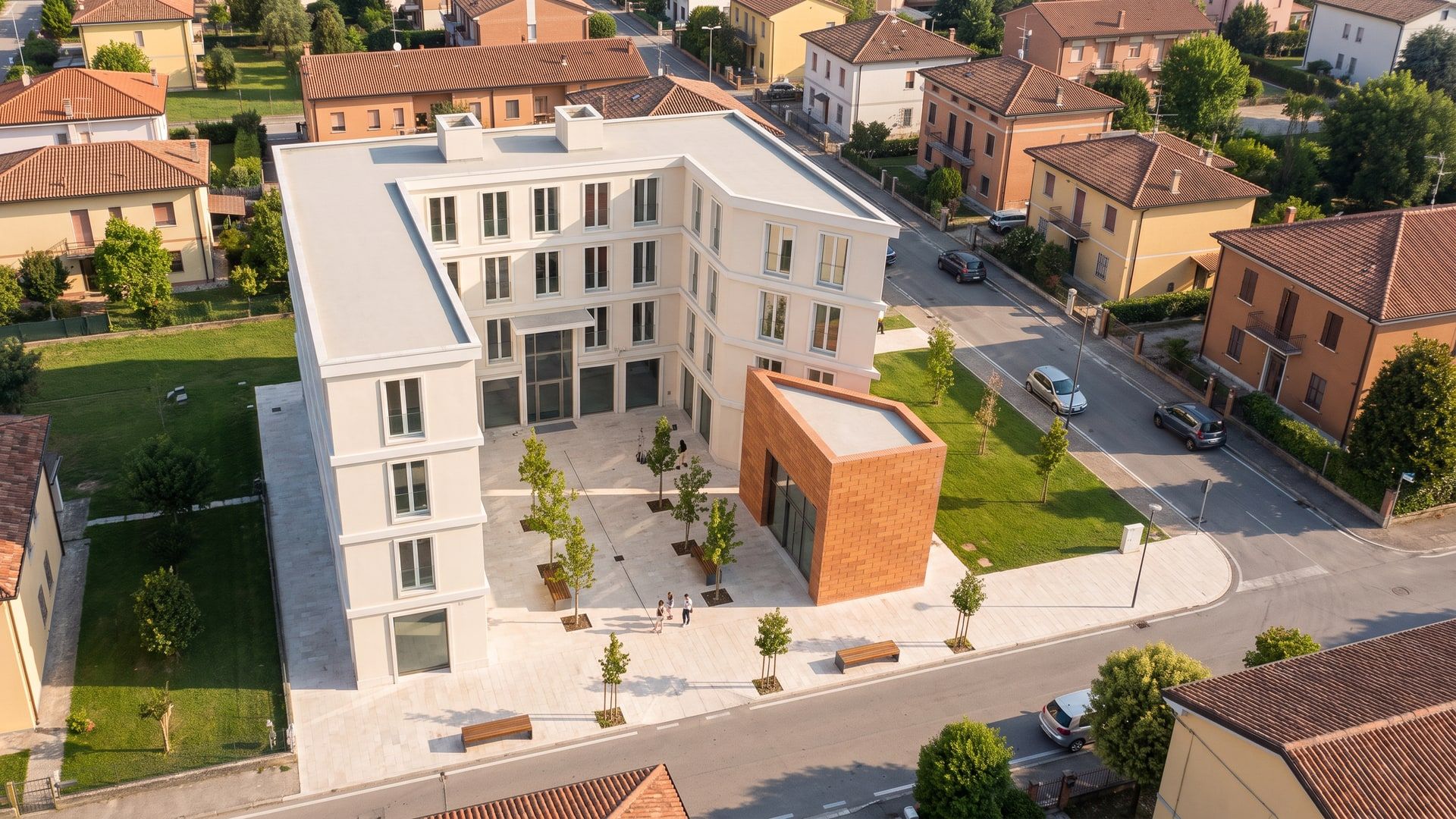 Render dall’alto: l’edificio bianco a corte con la piazza alberata e il padiglione in cotto, in un quartiere di case basse.